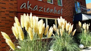Un bâtiment en briques avec une enseigne indiquant « Akazienhof » et des touffes d’herbe de la pampa devant.