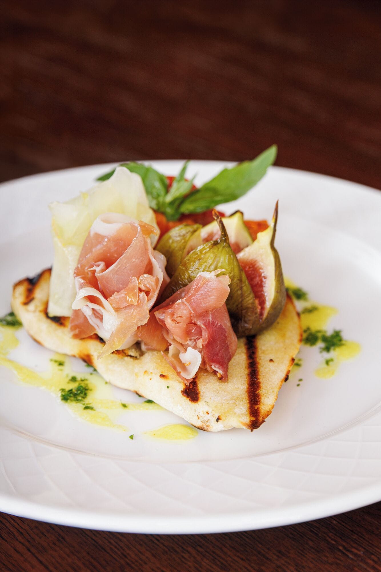 A plate with a grilled item, possibly a pancake or flatbread, topped with prosciutto and figs, garnished with herbs.