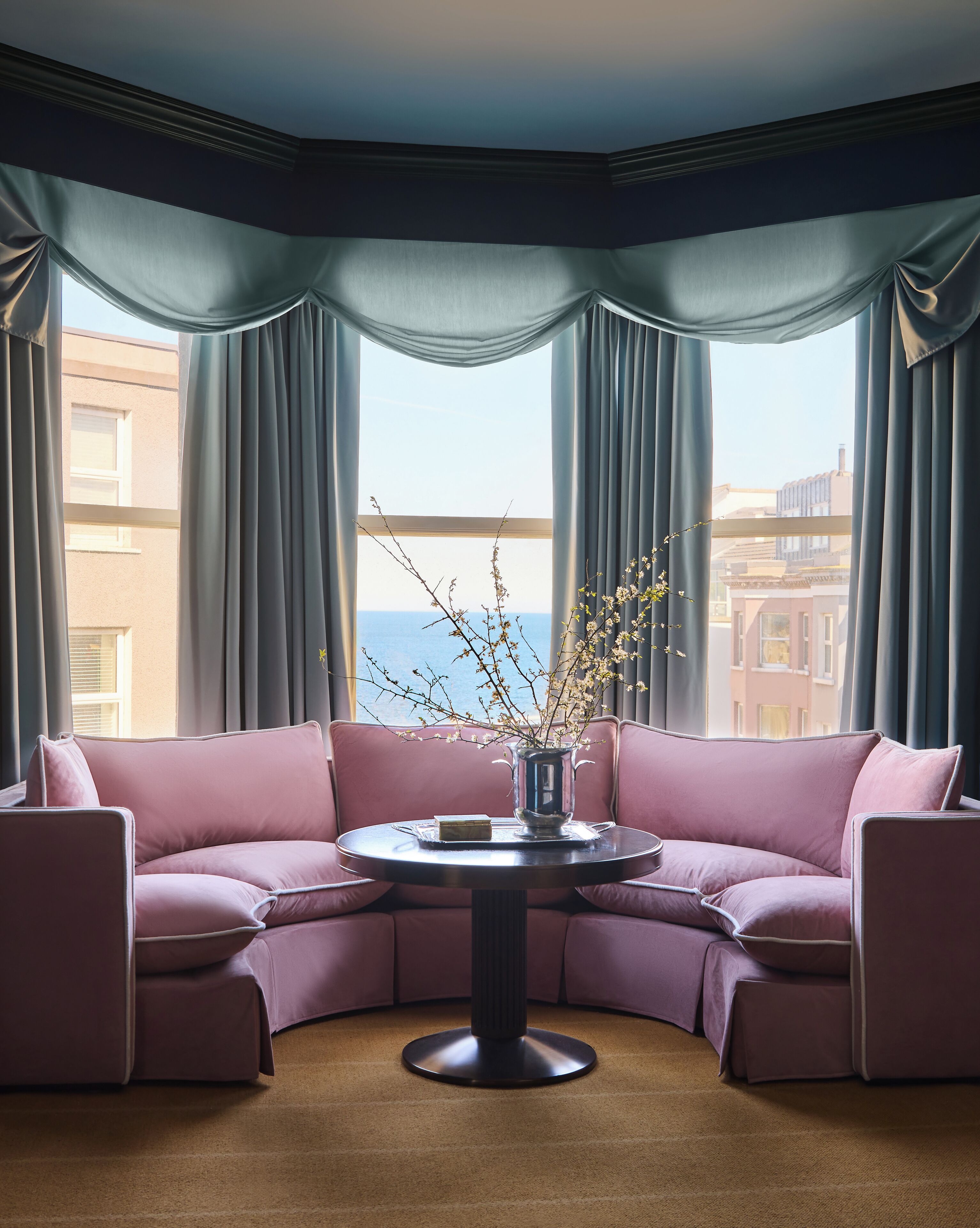 A corner sofa with a round table in between, a vase with branches, and a view of buildings through large windows.