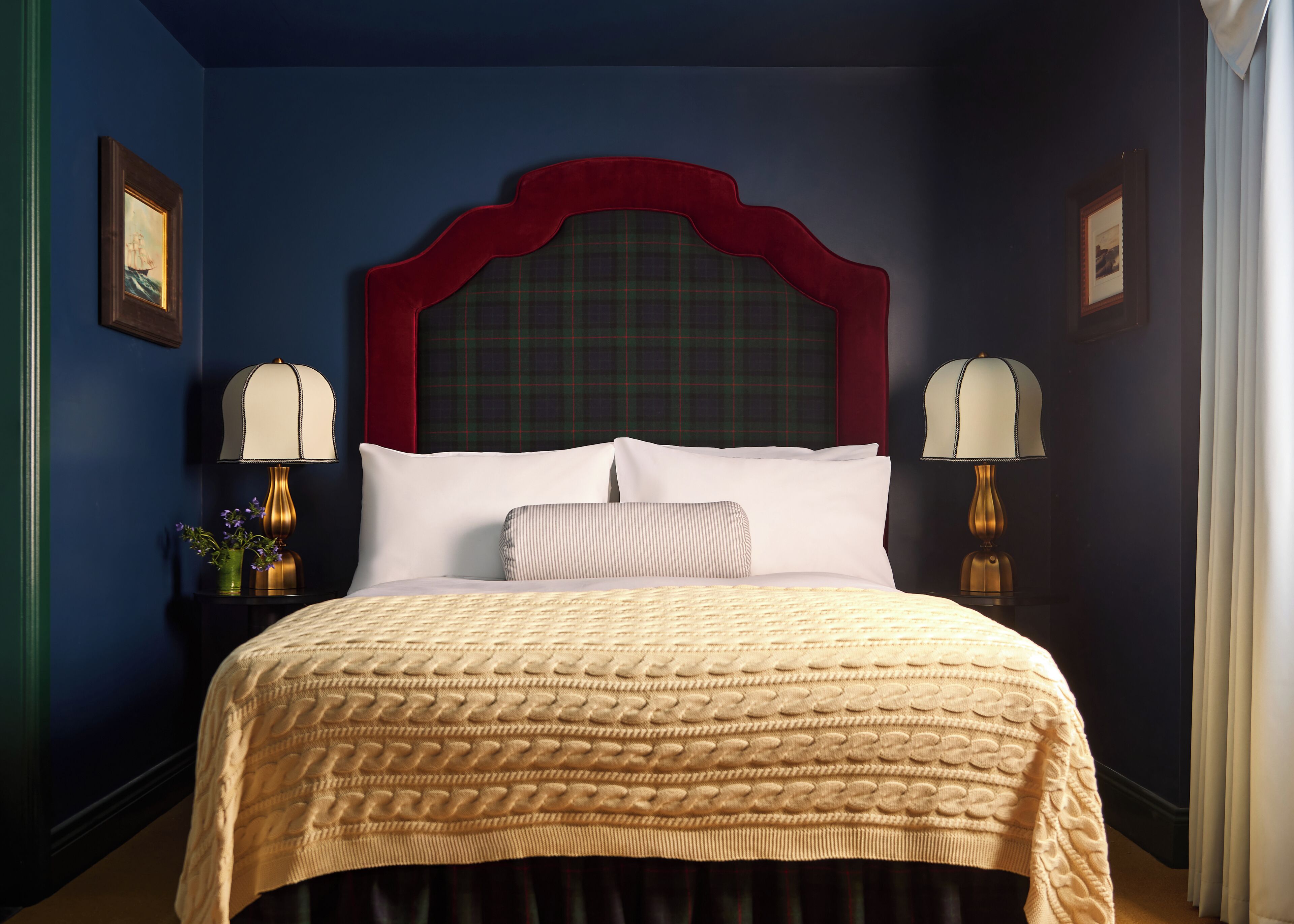 Quarto de hotel com uma cama grande, cabeceira vermelha, duas luminárias de apoio e cortina verde xadrez.