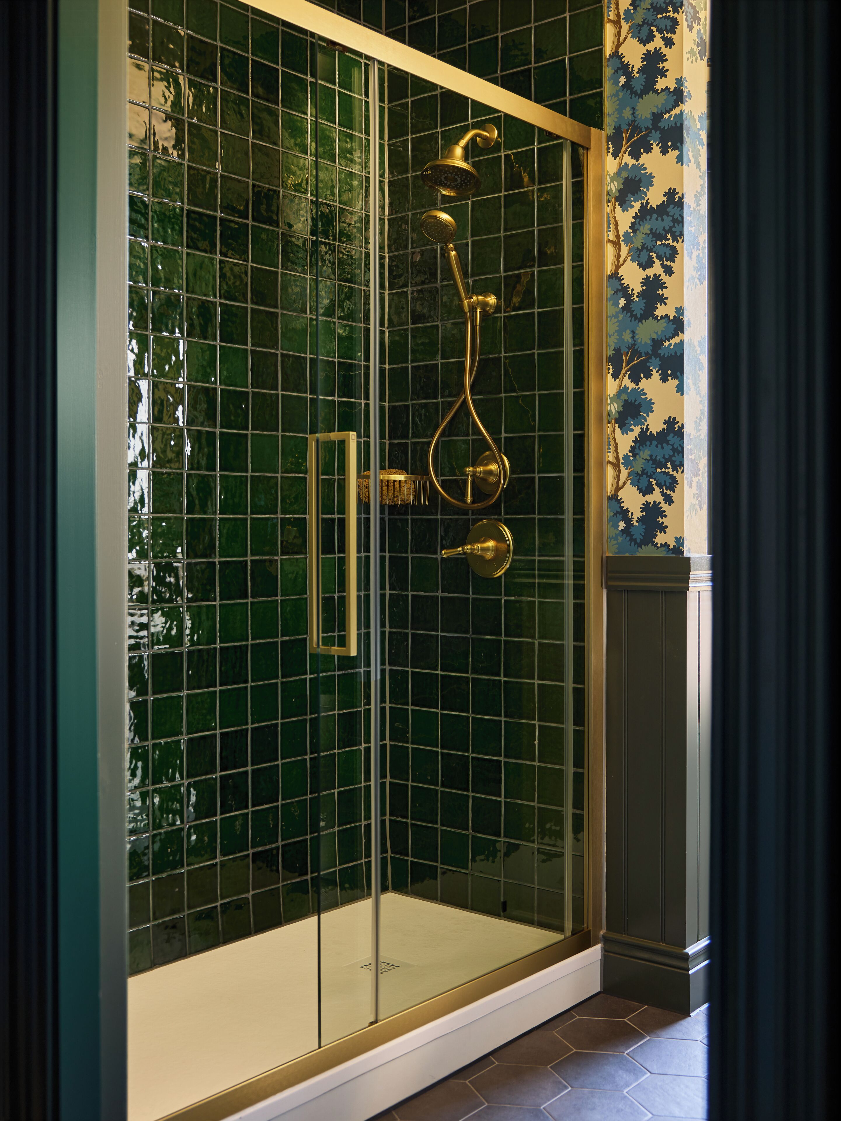A glass-enclosed shower with a gold showerhead and handle, set against a green-tiled wall.