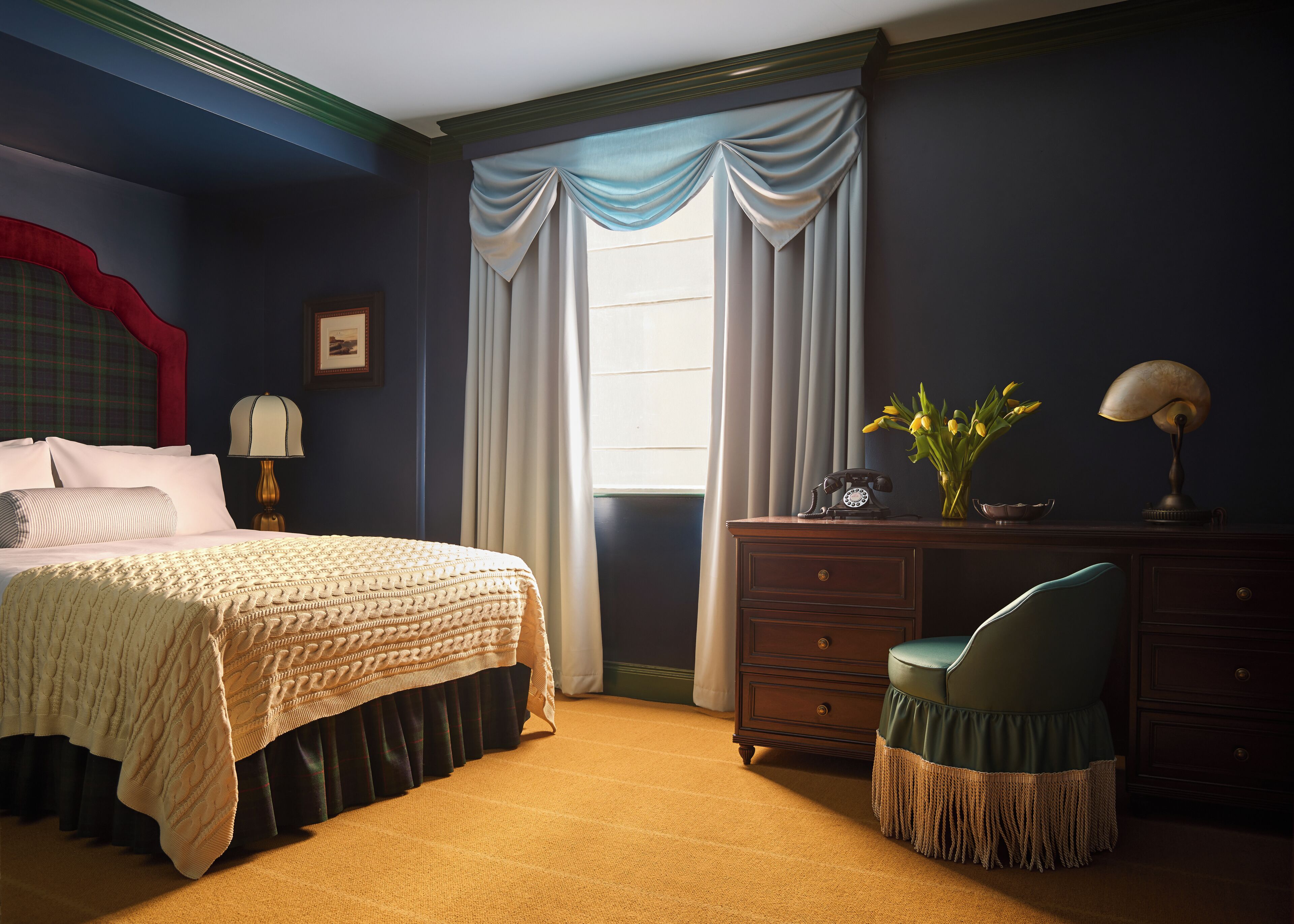 Une chambre à coucher comprenant un lit, une commode, un bureau avec une lampe, une chaise et une fenêtre avec des rideaux.