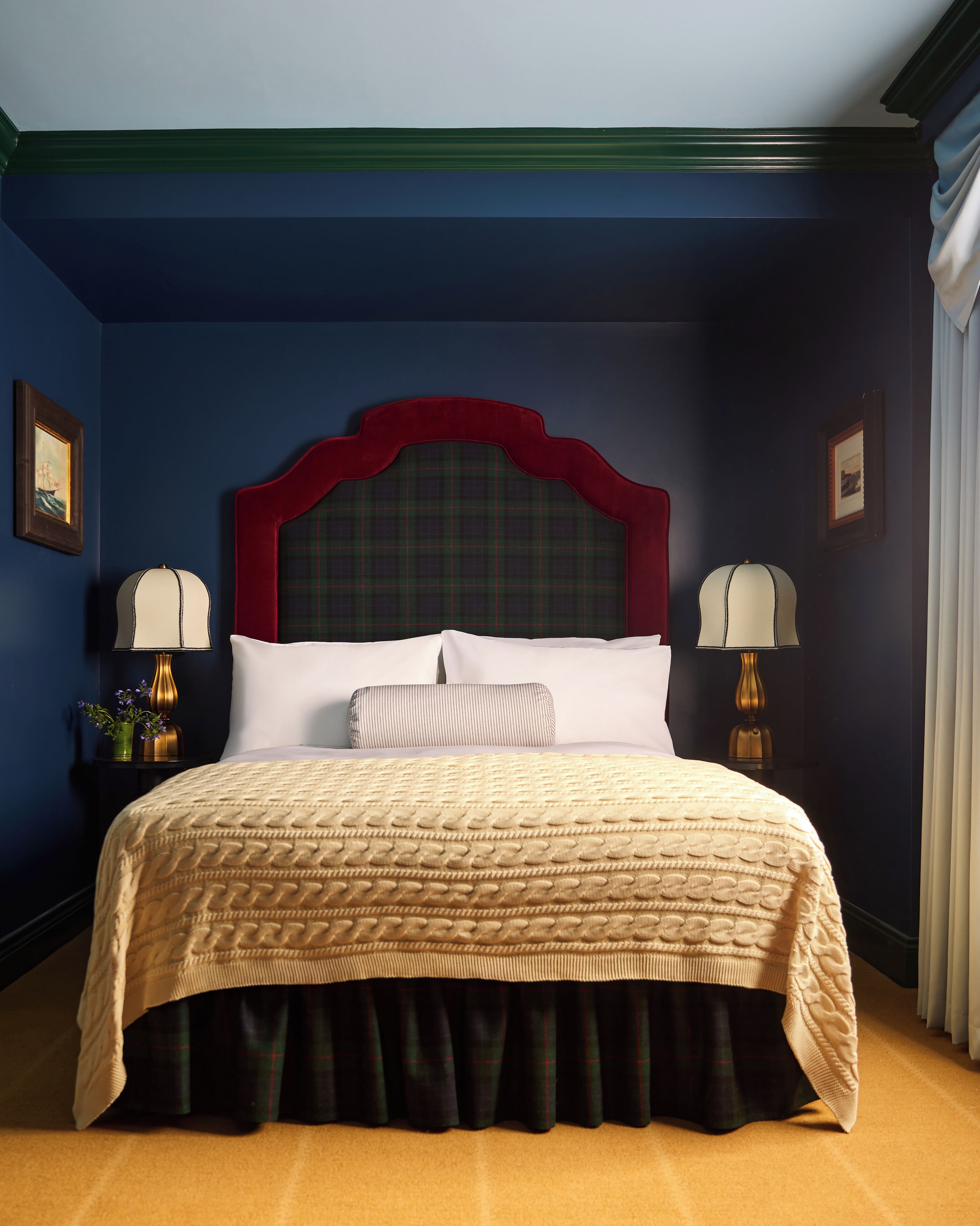A neatly made bed with a red headboard, two bedside lamps, and a plaid skirted bedspread.
