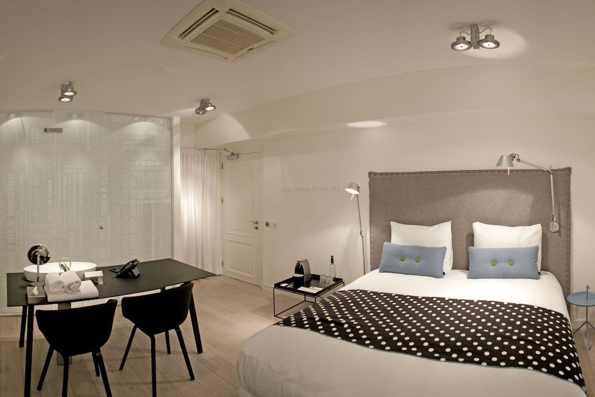 A modern hotel room with a bed, bedside table, lamp, and a desk with chairs.
