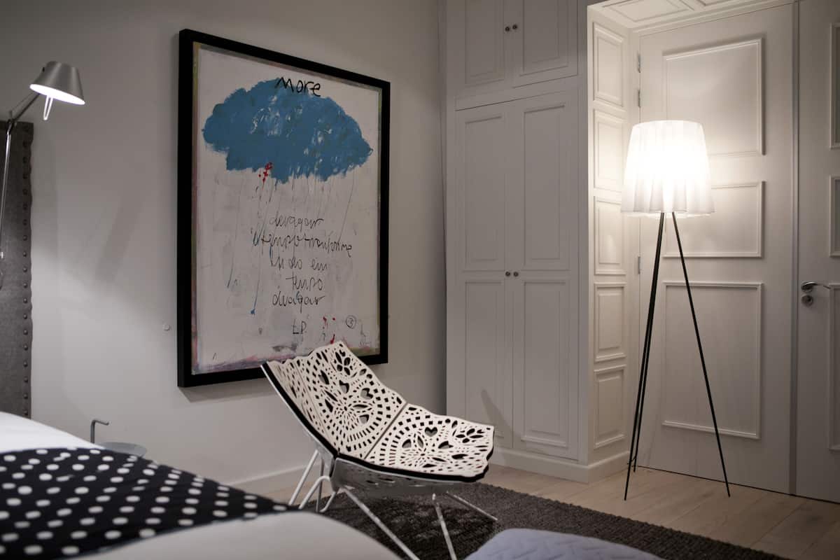 A room with a white chair, a framed artwork on the wall, a standing lamp, and a door.
