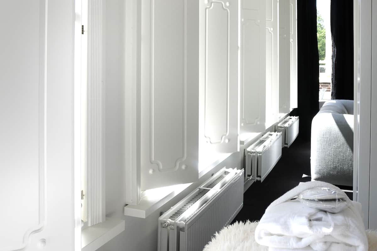 A hotel room with white walls, a radiator, a fluffy white rug, and a bed with a white cover.