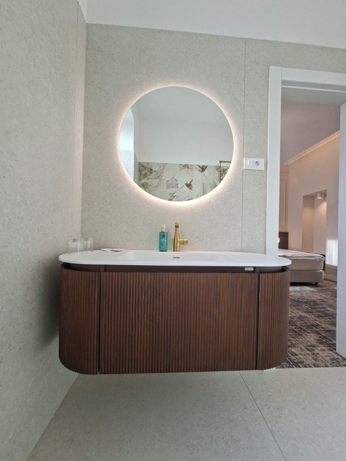 A modern bathroom with a freestanding vanity, a round mirror with integrated lighting, and a marble backsplash.
