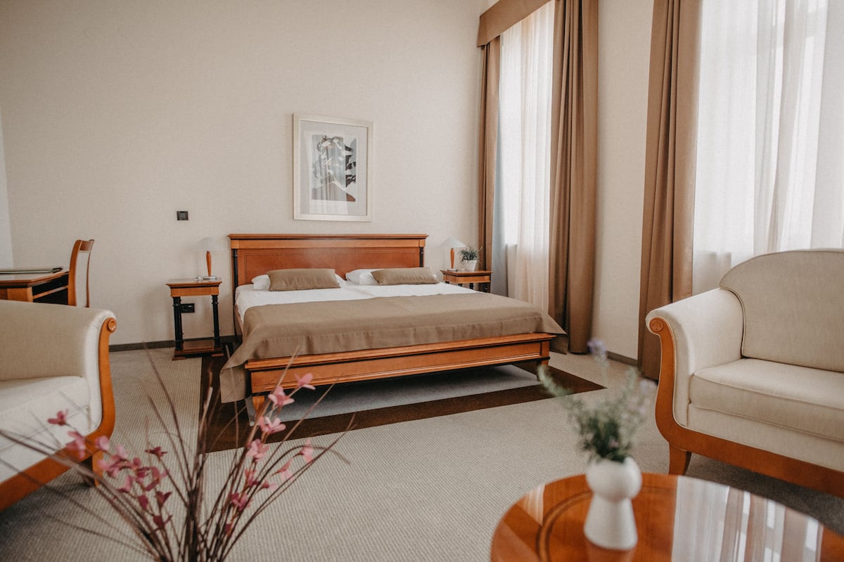 Una habitación de hotel con una cama, dos mesitas de noche, una silla, una mesita pequeña y un ventanal con cortinas.