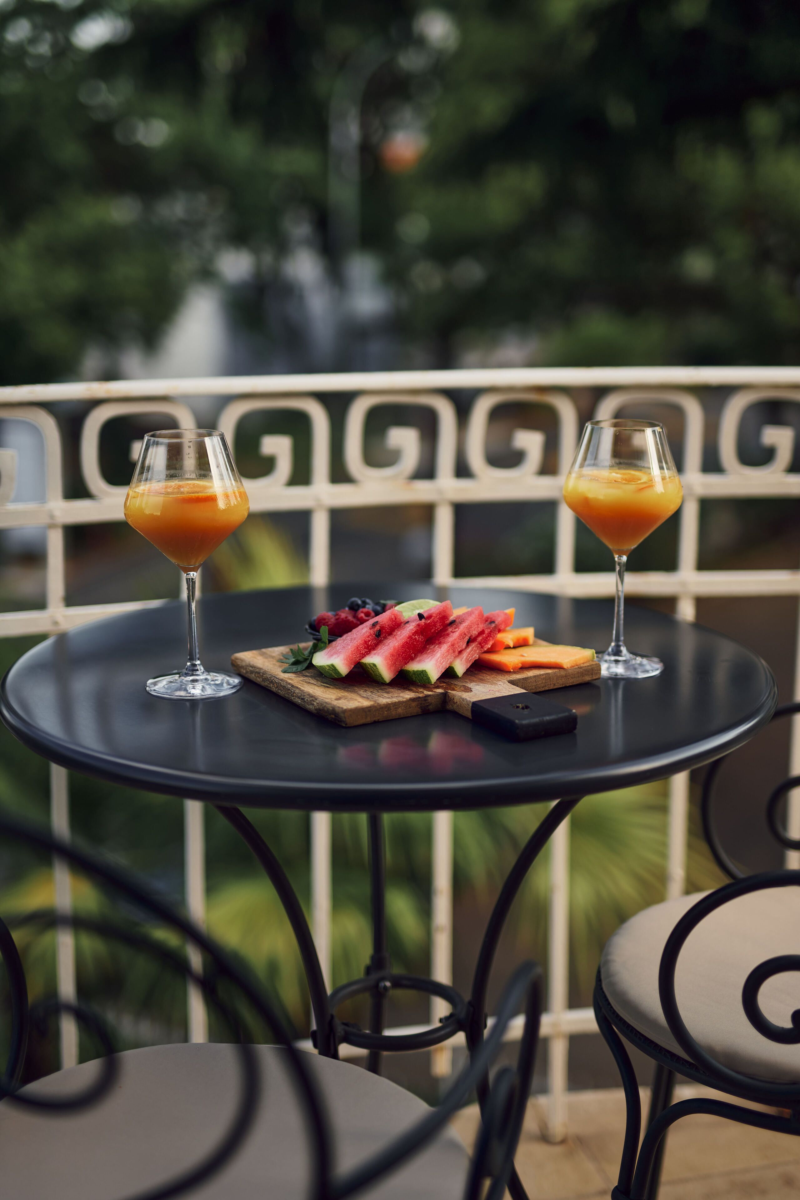 A table with two drinks and a fruit platter on a balcony with metal railings and greenery in the background.