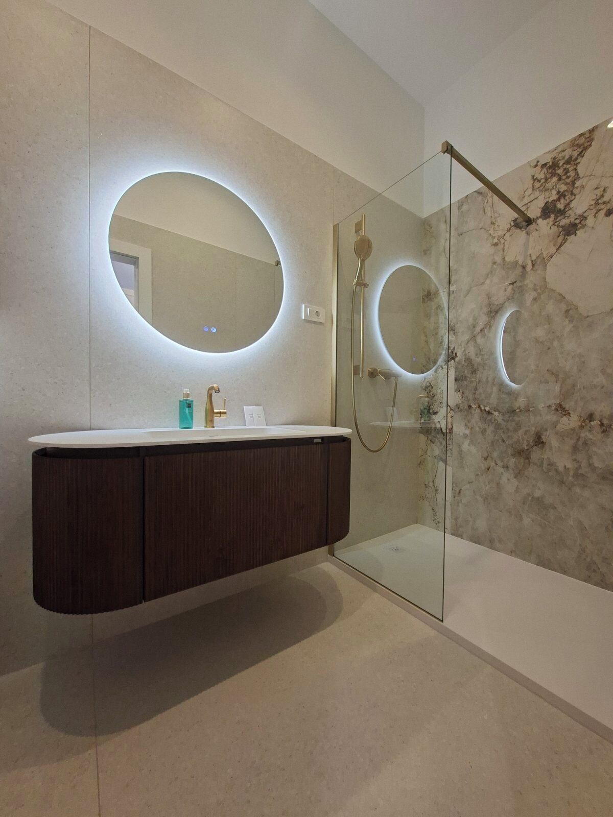 A modern bathroom with a marble wall, a glass-enclosed shower, and a dark wood vanity with a round mirror.