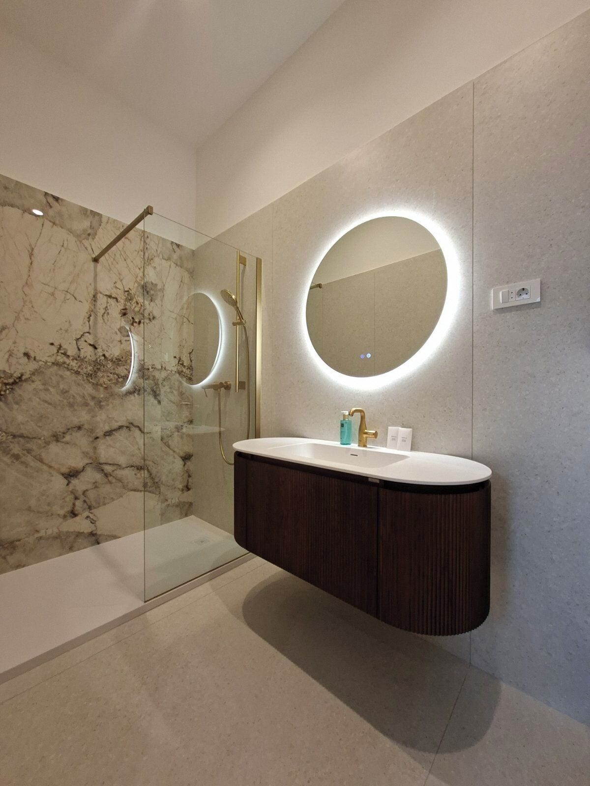 A modern bathroom with a marble shower, a vanity with a round mirror, and a built-in sink.