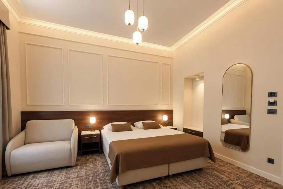 Una habitación de hotel con cama, una silla, mesitas de noche, un espejo y suelo alfombrado.