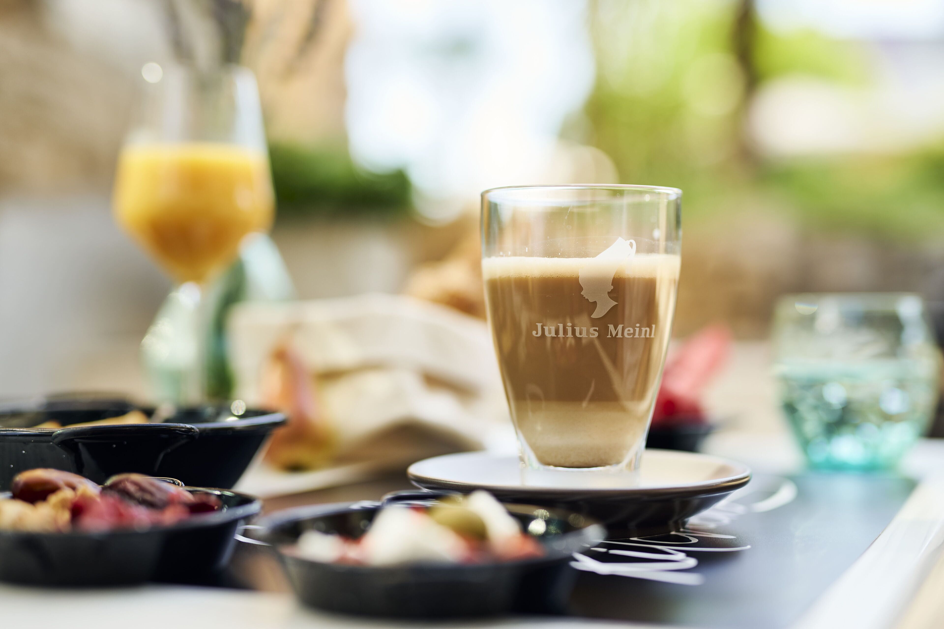 Eine Tasse Kaffee mit Untertasse auf einem Tisch mit verschiedenen Speisen und im Hintergrund ein Glas Orangensaft.