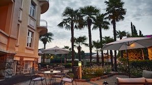 An outdoor patio area with tables and chairs, umbrellas, and palm trees. - Hotel Villa Eugenia (Lovran)