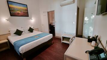 A hotel room with a bed, bedside table, desk with a phone, and an air conditioning unit.