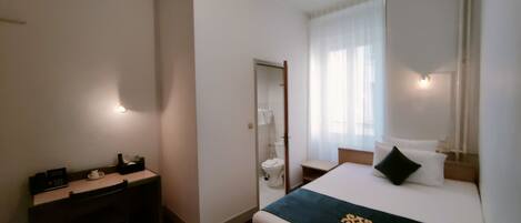 A hotel room with a bed, bedside table, and a small desk with a chair. There is a window with curtains and a door leading to a bathroom.