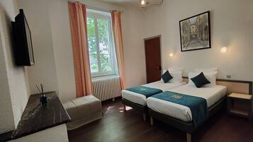 Une chambre d’hôtel avec deux lits, une fenêtre avec des rideaux, un radiateur, un téléviseur fixé au mur et un tableau accroché au mur.