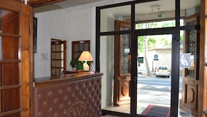 A hotel reception area with a wooden reception desk, a lamp, and a view of the street outside. - Will's Hotel (Narbonne)