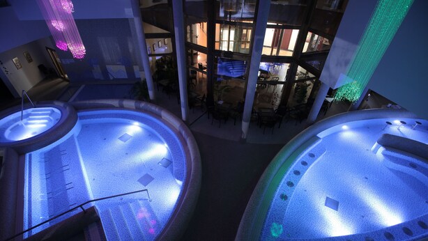 A multi-level indoor space with a central atrium, multiple seating areas, and a pool with illuminated edges. - Colosseum Hotel Mórahalom (Morahalom)