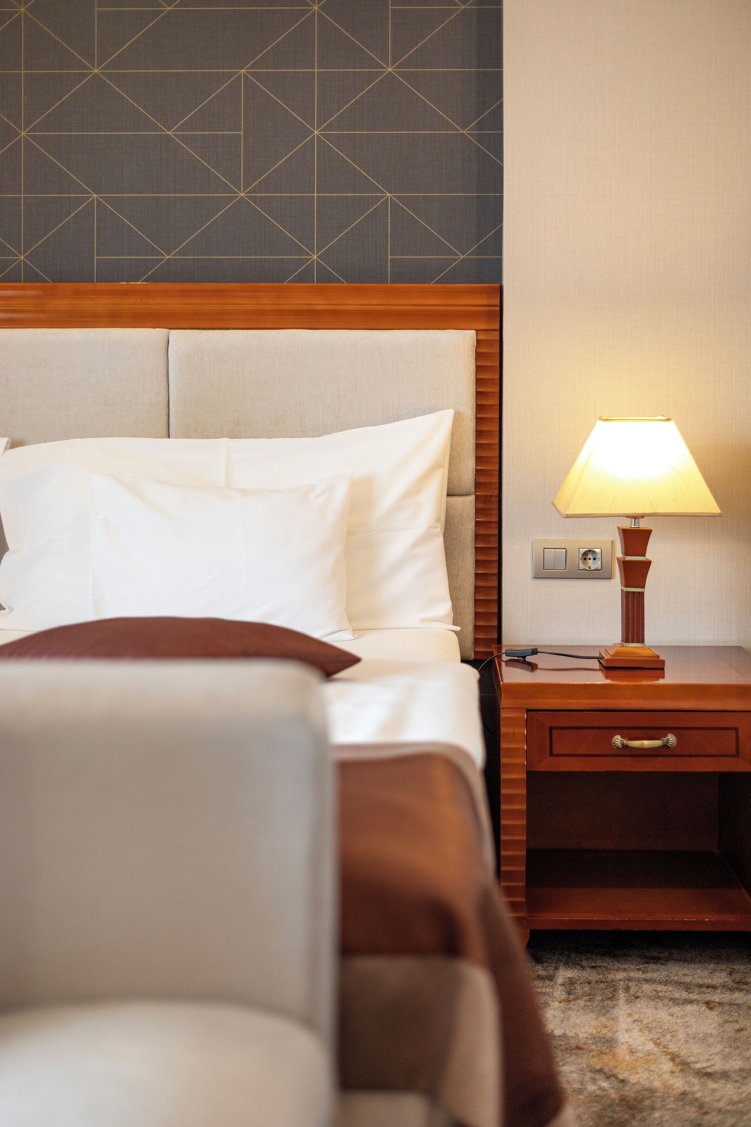 A hotel room with a bed, a nightstand, a lamp, and a wall with a geometric pattern.