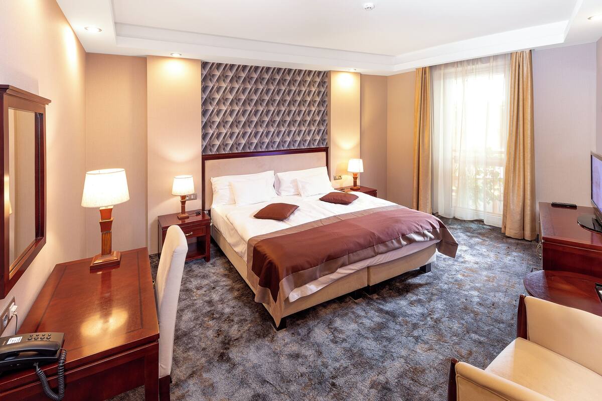 A hotel room with a large bed, bedside lamps, a desk with a telephone, and a television.