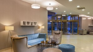 A lobby with a blue sofa, two armchairs, a coffee table, and a round ottoman. There are ceiling lights and a glass door leading outside. - Colosseum Hotel Mórahalom (Morahalom)