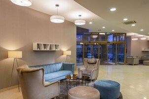 Un hall con un sofá azul, dos sillones, una mesa de centro y un reposapiés redondo. Hay luces en el techo y una puerta de cristal que da al exterior.