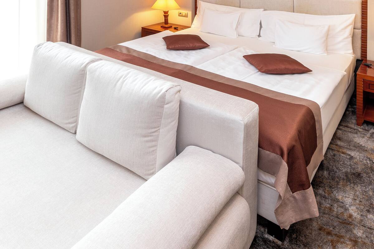 A hotel room with a large bed, a sofa, a nightstand with a lamp, and a wall with a geometric pattern.