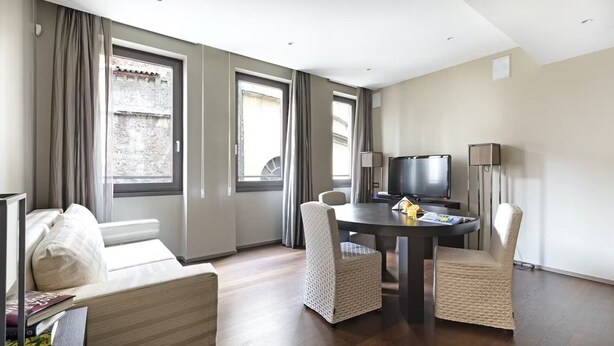Ett modernt hotellrum med en soffa, ett matbord med stolar, en TV och stora fönster med gardiner.