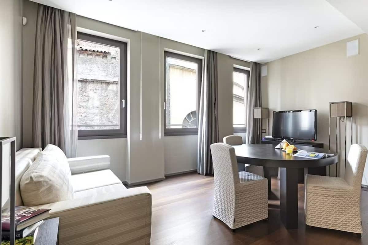 A modern hotel room with a sofa, a dining table with chairs, a TV, and large windows with curtains.