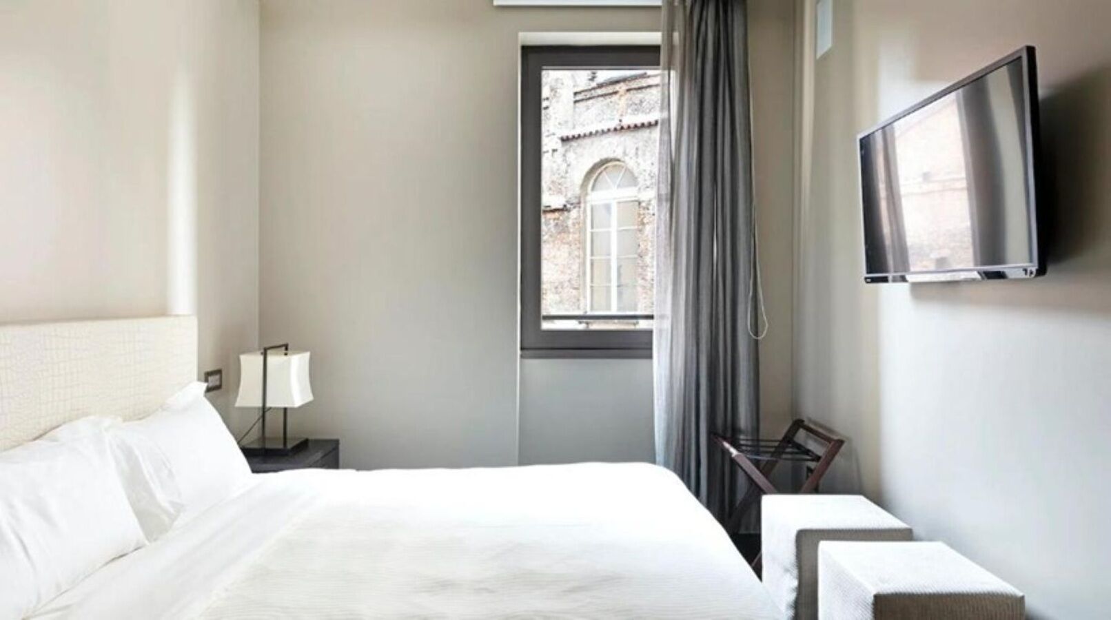 Ein Hotelzimmer mit Bett, einem an der Wand befestigten Fernseher, einem Fenster mit Vorhängen und Blick auf ein Gebäude.