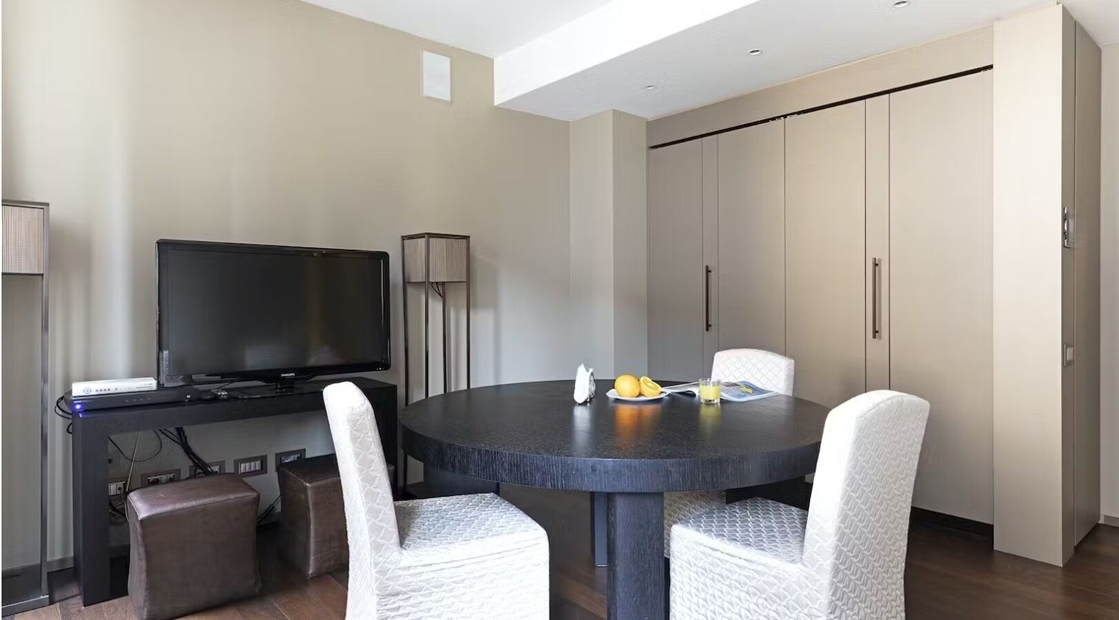 Een moderne hotelkamer met een ronde eettafel, twee witte fauteuils, een flatscreen televisie, een minibar en een grote inbouwkast.