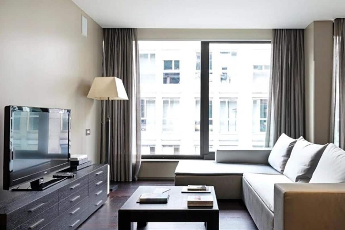 A modern hotel room with a flat-screen TV, a sofa, a coffee table, and a large window with curtains.
