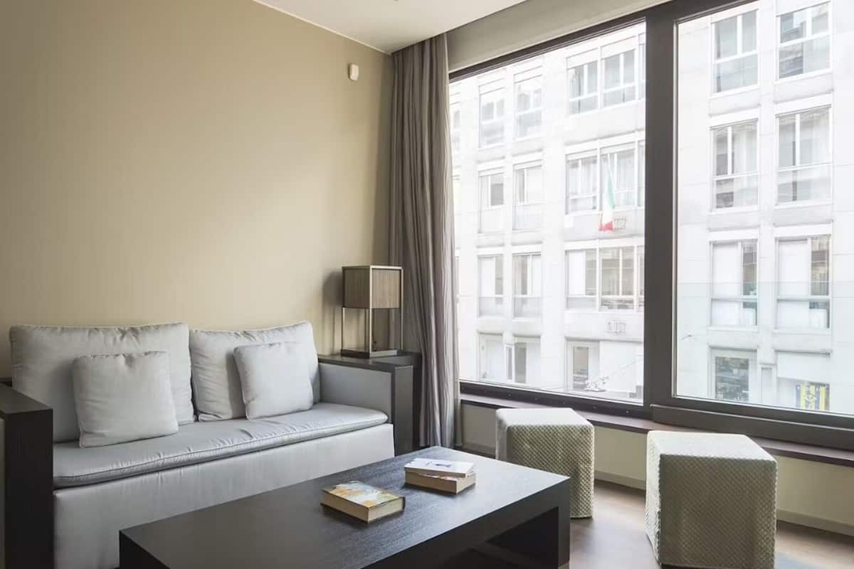 A modern hotel room with a sofa, a small table, a lamp, and a view of a building through large windows.