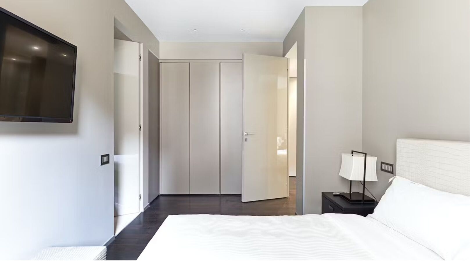 A hotel room with a bed, a TV mounted on the wall, a bedside table with a lamp, and a door leading to another room.