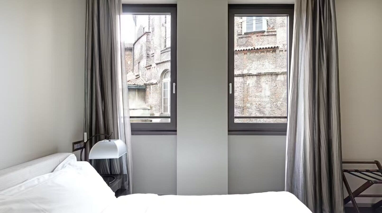 A hotel room with a bed, a desk lamp, and two windows with curtains.