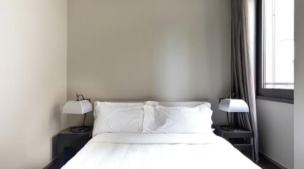 A hotel room with a bed, two bedside lamps, a window with curtains, and a plain wall.
