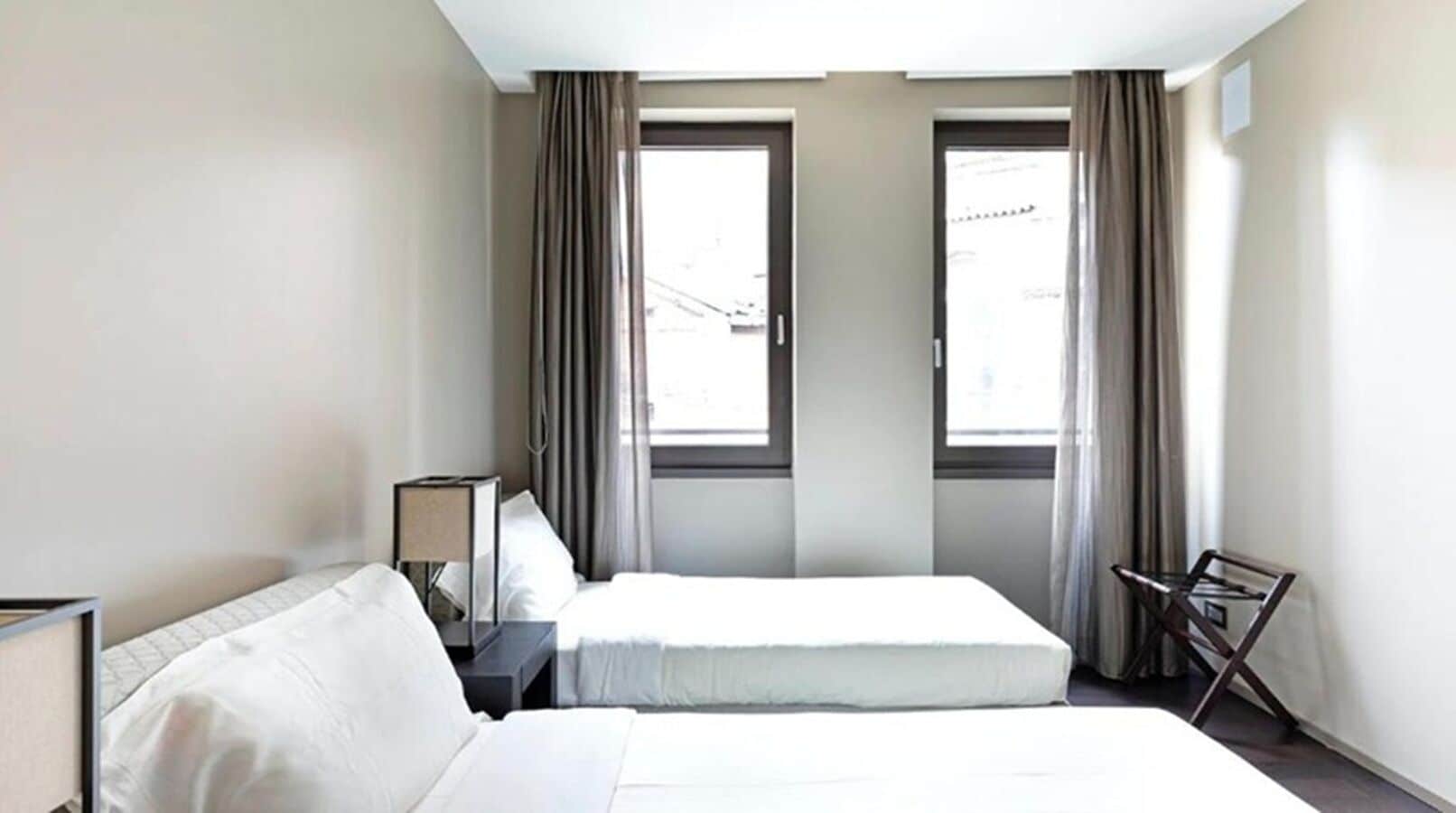 A hotel room with two beds, a desk, a chair, and two windows with curtains.