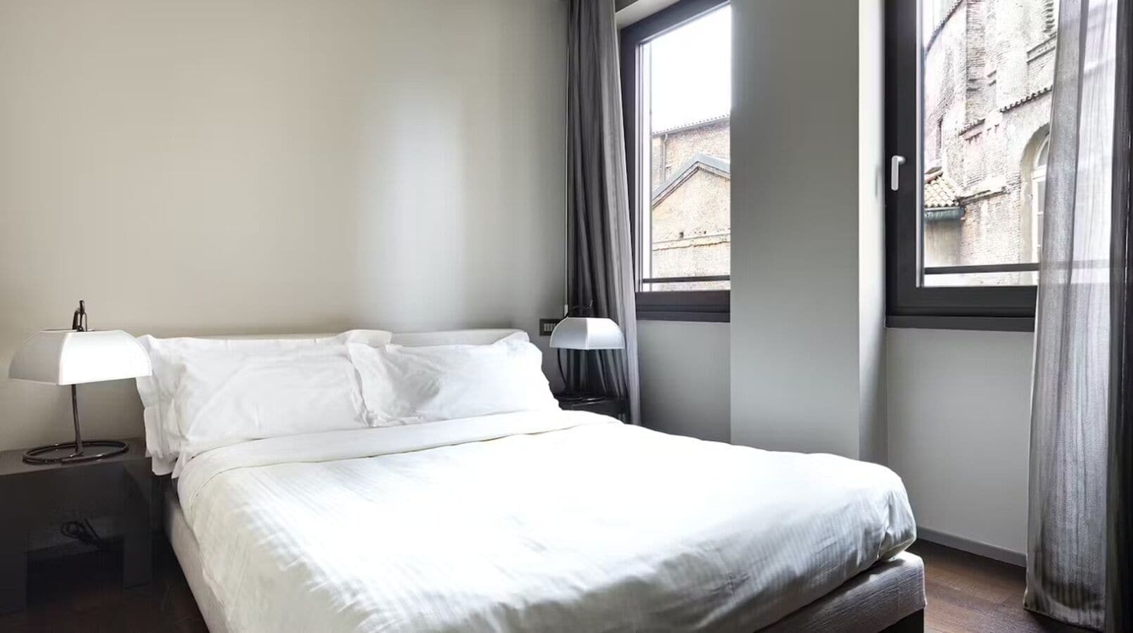 A neatly made bed in a hotel room with a window, a bedside lamp, and a view of a building outside.