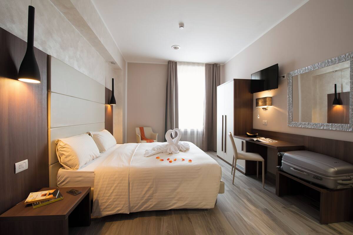 A hotel room with a bed, bedside table, desk, chair, and a suitcase.