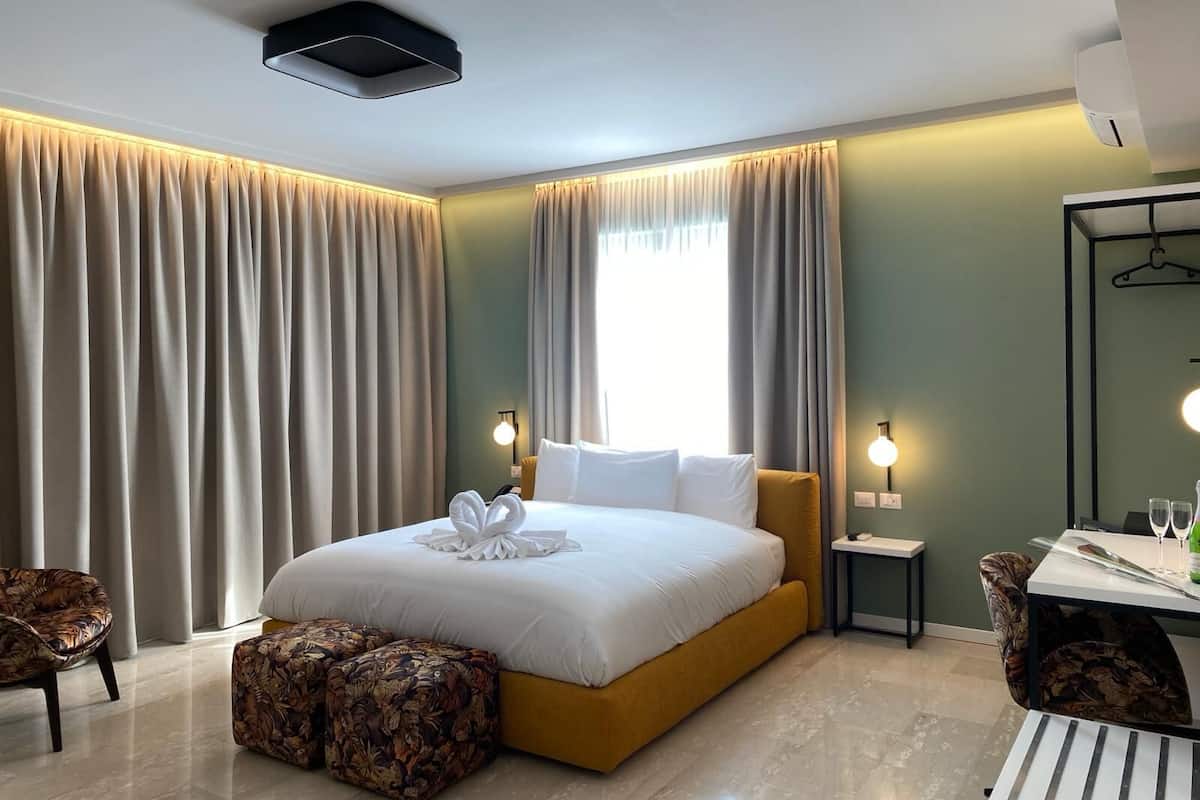 A modern hotel room with a large bed, a small table with two chairs, a mirror, and a window with curtains.