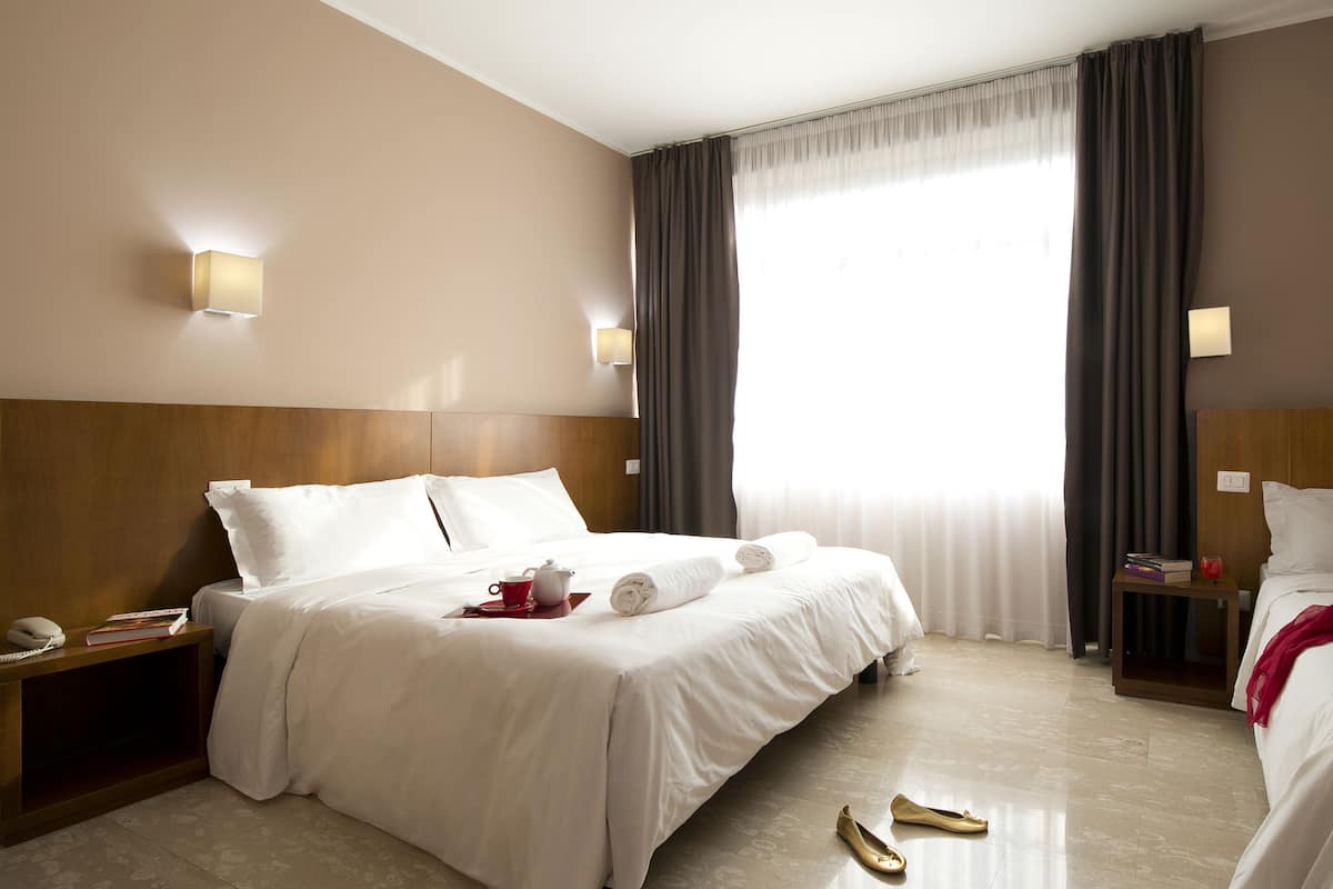 A hotel room with two beds, a bedside table with a lamp, a nightstand, a window with curtains, and a wooden headboard.
