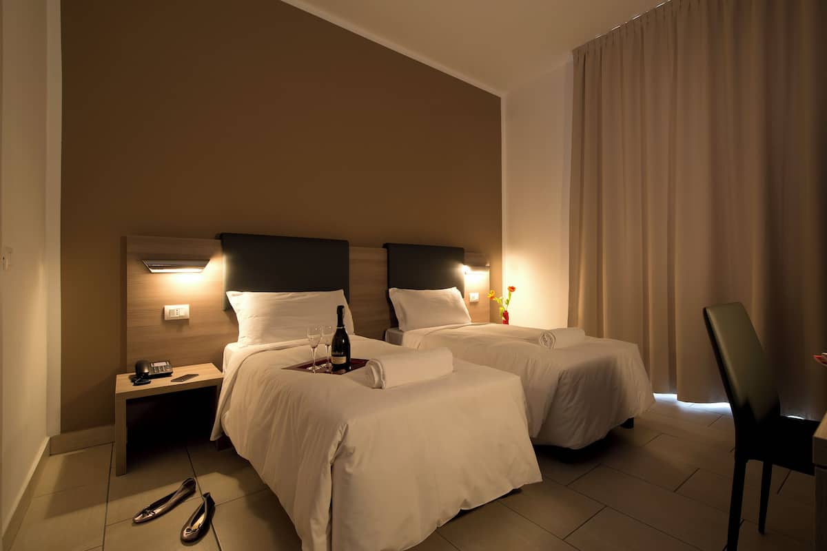 A hotel room with two beds, a desk with a chair, a telephone, and a bottle of champagne on the nightstand.