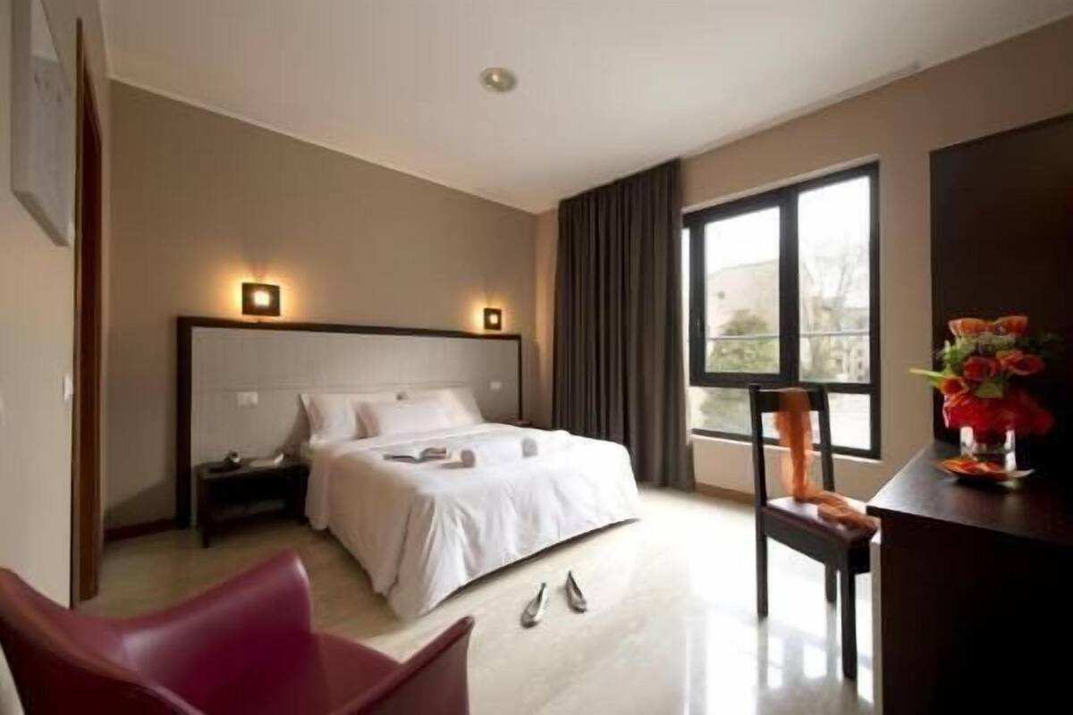 A hotel room with a bed, bedside tables, a desk with a chair, a window with curtains, and a vase with flowers.