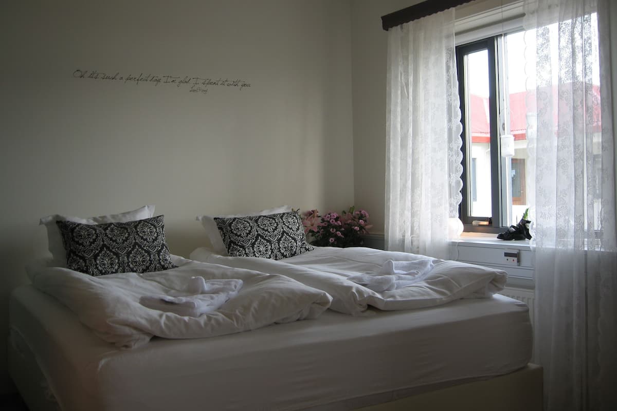 A bedroom with two beds, white bedding, patterned pillows, a window with sheer curtains, and a wall with a handwritten message.