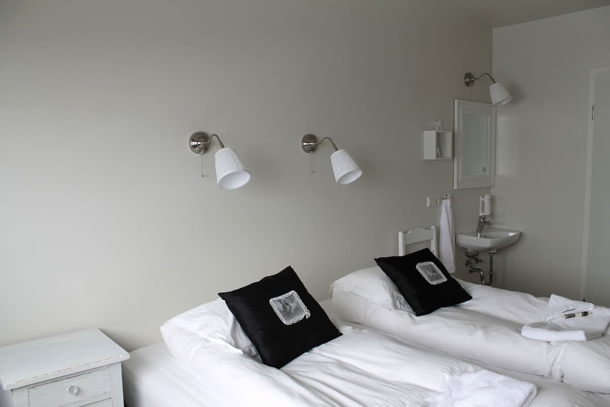A hotel room with two beds, a nightstand, a white dresser, a wall-mounted sink, and two wall sconces.