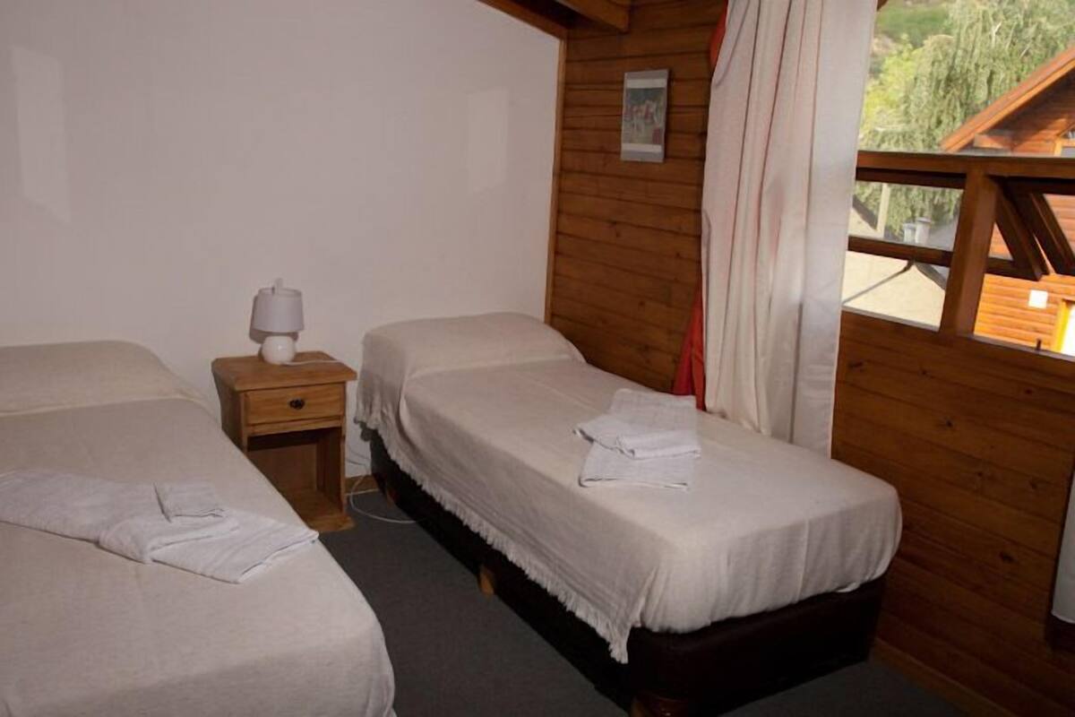 A room with two single beds, wooden walls, a small bedside table with a lamp, and a window with curtains.