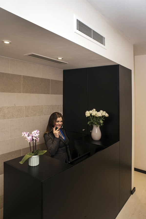 A reception desk with a vase of white flowers, a potted plant, and a person standing behind it. - NOX HOTELS - Notting Hill (London)