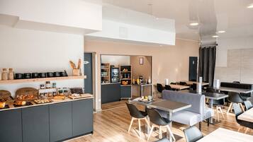 Un café moderno con un mostrador central que exhibe pasteles y bebidas, zonas de estar con mesas y sillas, y una cocina al fondo.