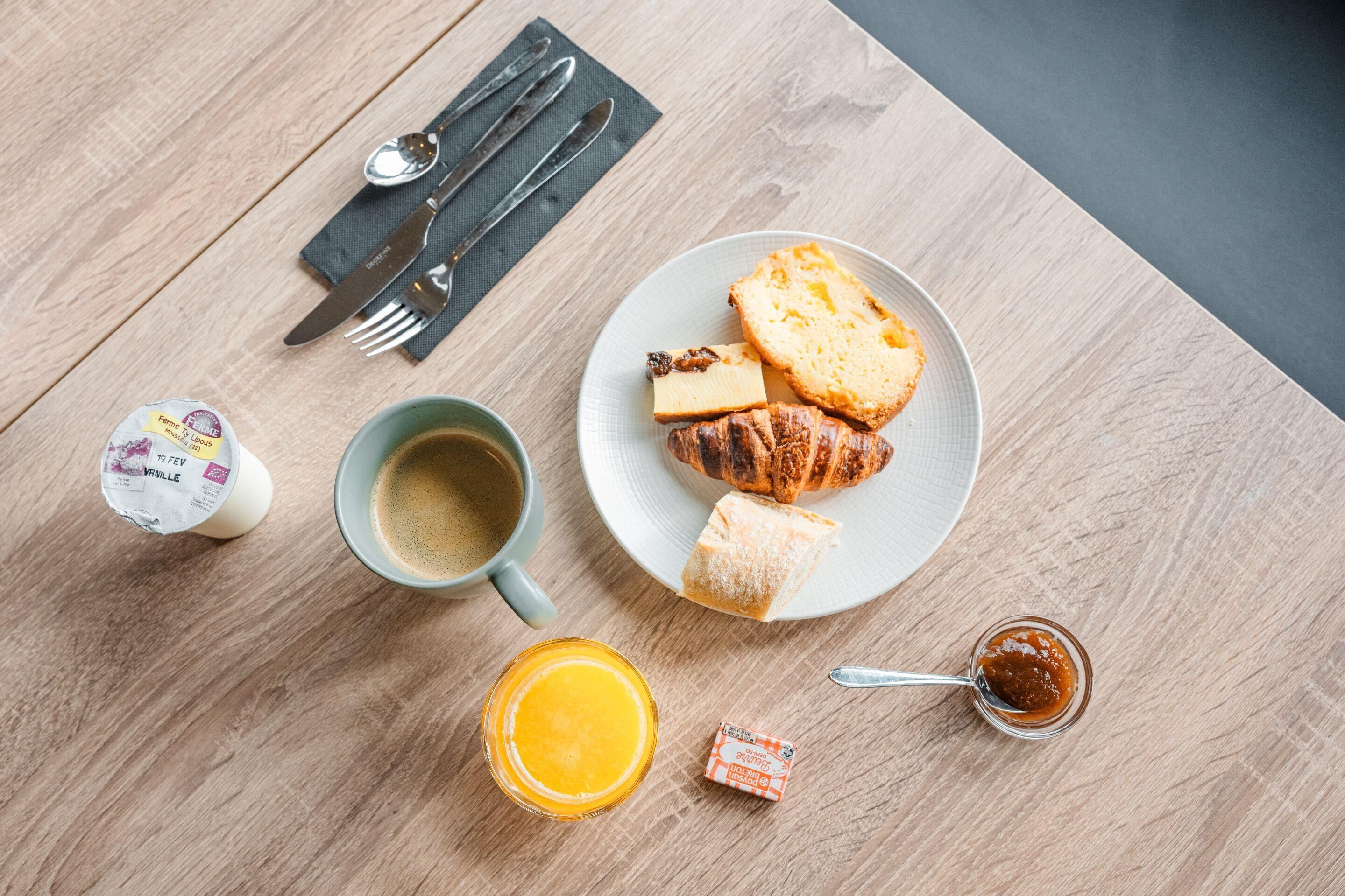 Un desayuno con un croissant, queso, mermelada en un plato, una taza de café, un frasco pequeño de mermelada y un envase de yogur.